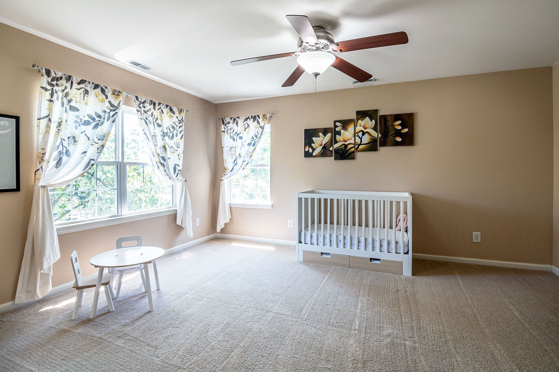 A spacious nursery room with a crib, white table and chair set, and floral curtains under natural light.