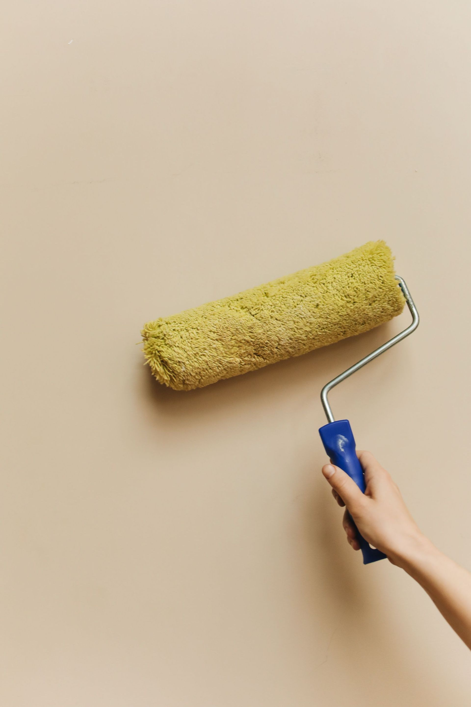 Hand holding a paint roller applying fresh coat on beige wall in home improvement project.